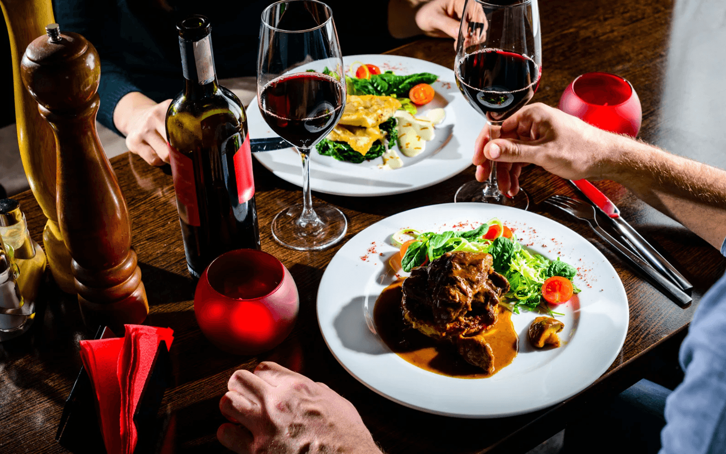 A table setting featuring two plates of gourmet meals, red wine glasses, and a bottle of red wine.