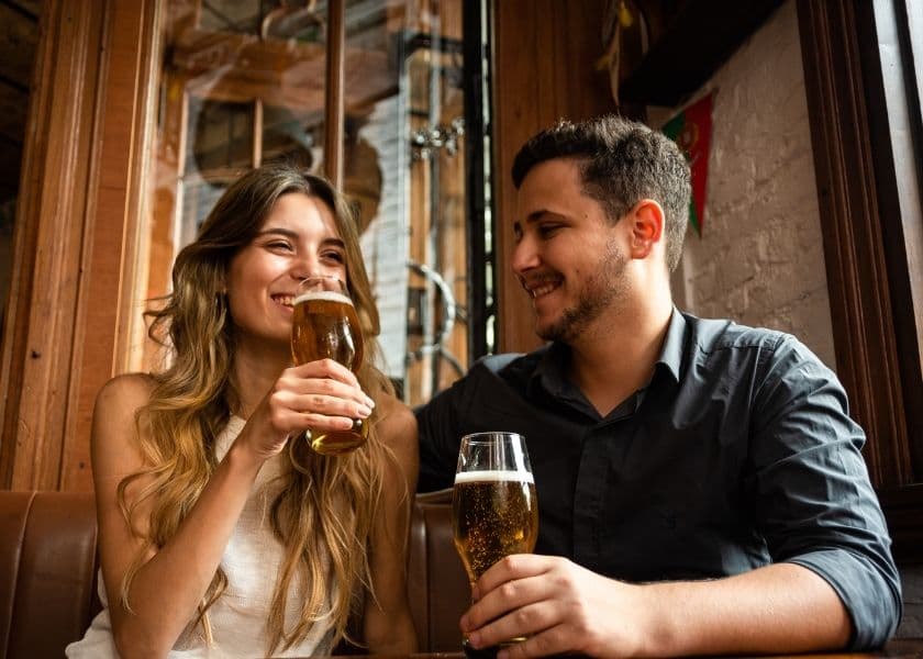 A smiling couple clinks glasses of beer together in a cozy bar setting. A smiling couple clinks glasses of beer together in a cozy bar setting.