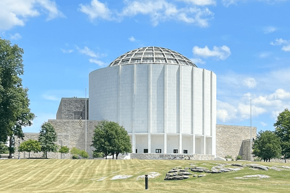 A modern circular building with a dome, set against a blue sky and green lawn.