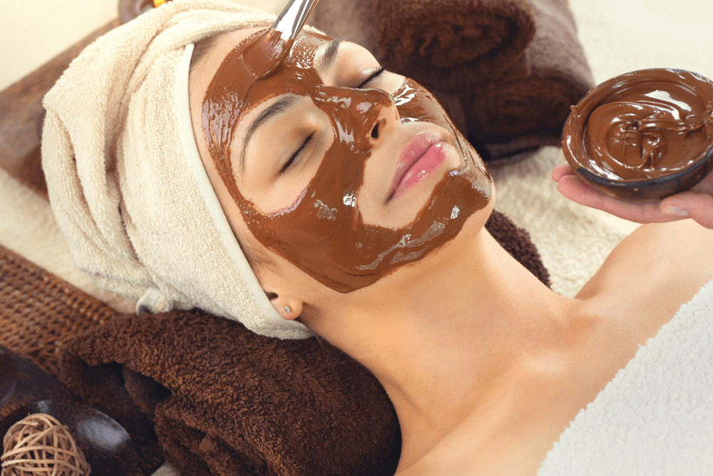 A woman relaxes with a chocolate facial mask while lying on a spa table.