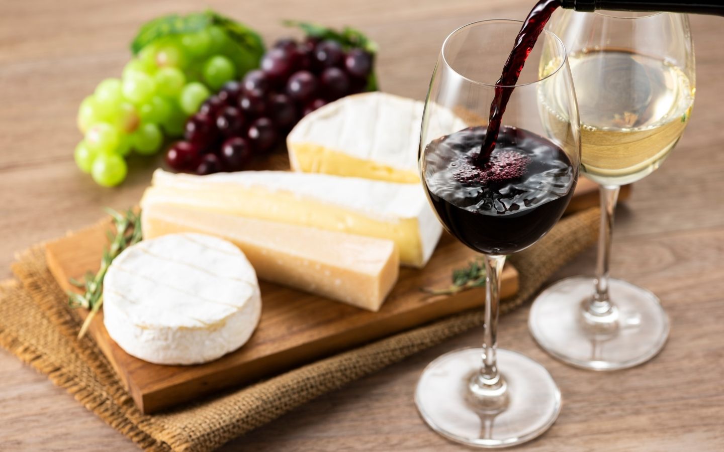 wooden board with assorted cheese and grapes next to a glass of white wine and glass of red wine with wine pouring into it